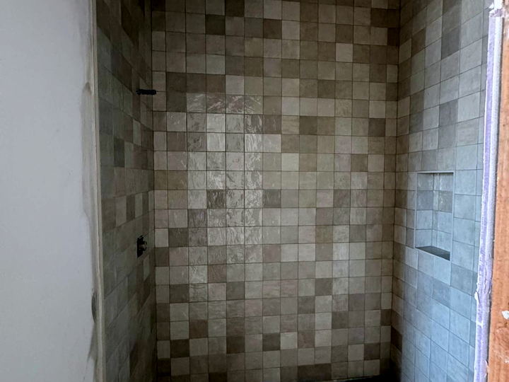 Shower walls tiled in varied beige and grey square tile with a recessed niche, before the pan is finished