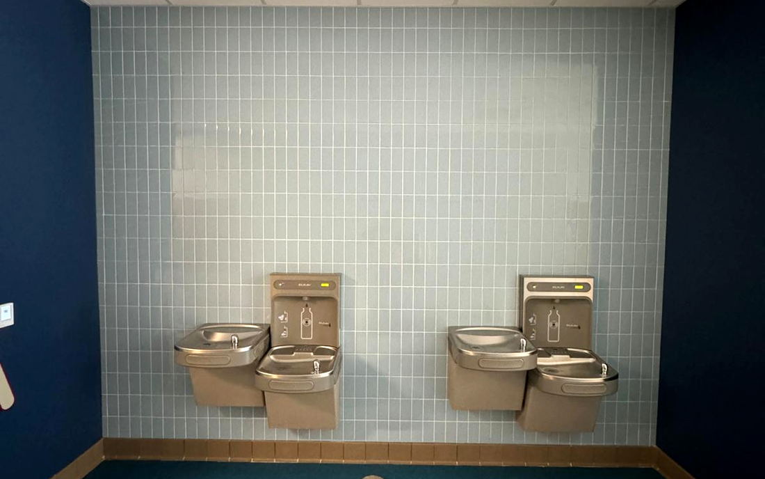 Pale blue tiled wall with two double drinking fountains, framed by navy walls and resilient flooring