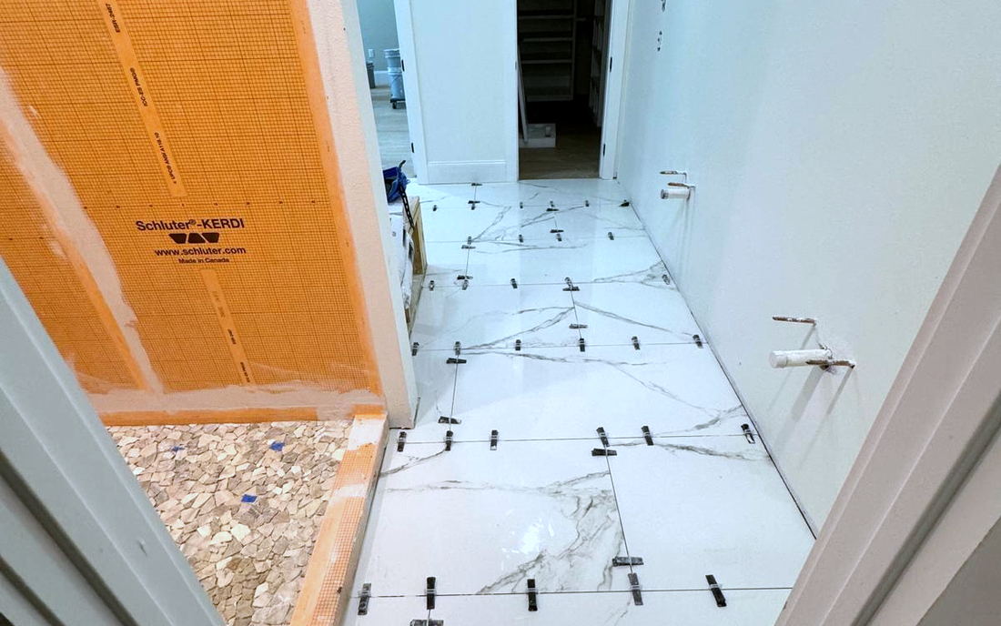 Shower wall covered in orange Schluter KERDI waterproofing membrane beside a marble-look porcelain floor being set with leveling clips