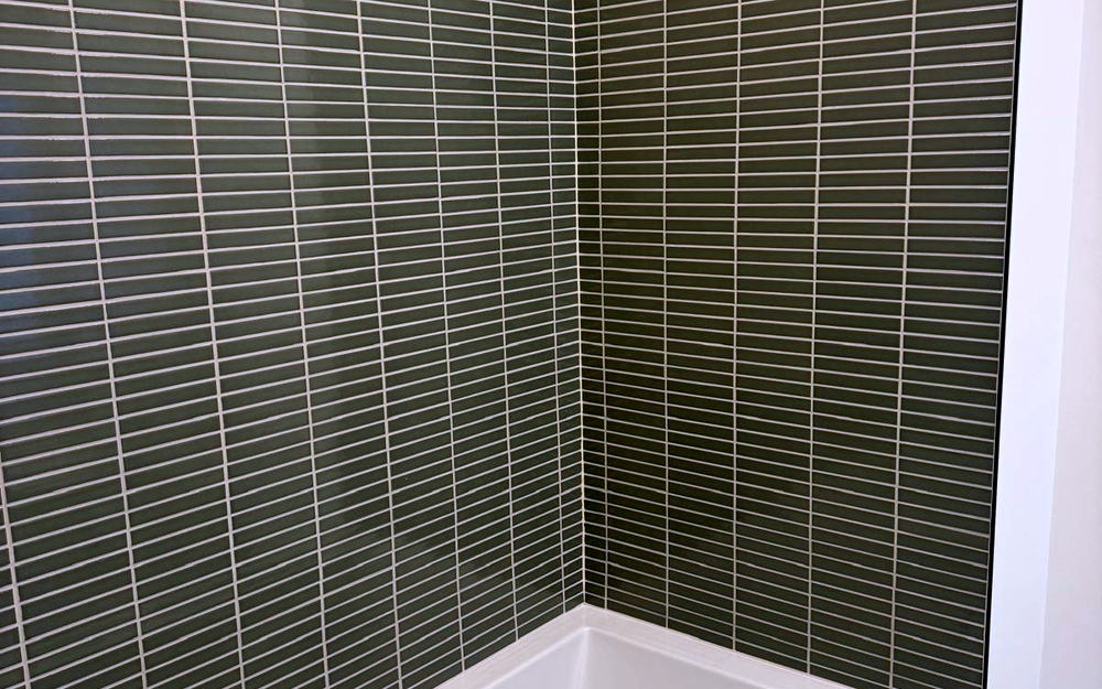 Tub surround in sage green stacked finger tile, mitered at the corner, above a white tub and a penny mosaic floor