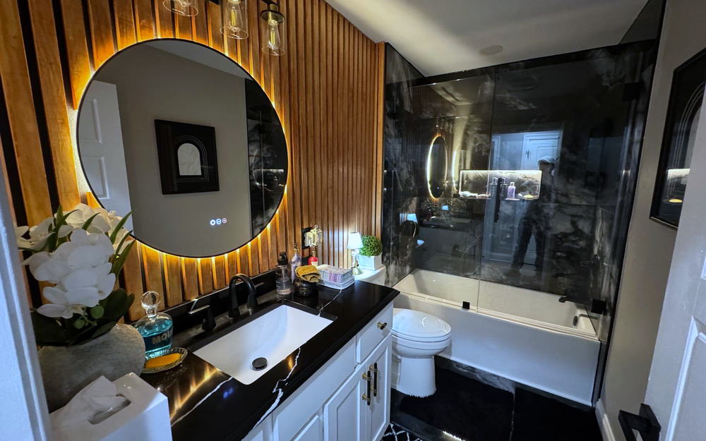 Full view of a dark bathroom with a slatted wood wall, black marble-look shower surround and a glass tub screen