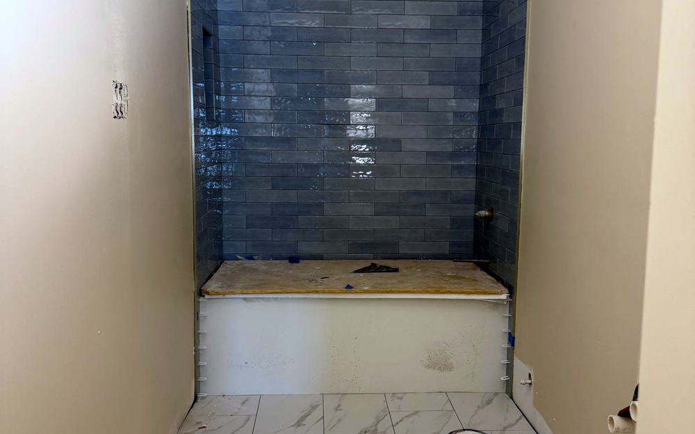 Tub alcove tiled in blue-grey subway to the ceiling, with a marble-look porcelain floor and the tub not yet set