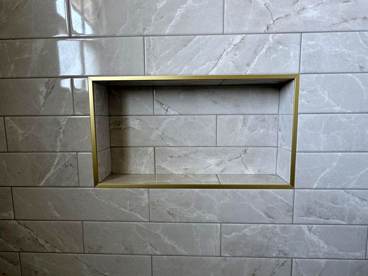 Recessed shower niche framed in brushed gold trim, set into white marble-look subway tile