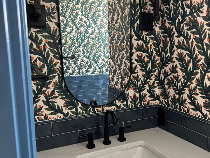 Vanity detail with a black framed oval mirror, wall sconces, botanical wallpaper and a dark blue tile backsplash