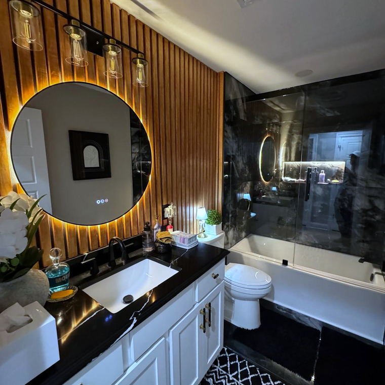 Bathroom vanity with a black marble-look countertop and backsplash against a slatted wood wall, with a backlit round mirror
