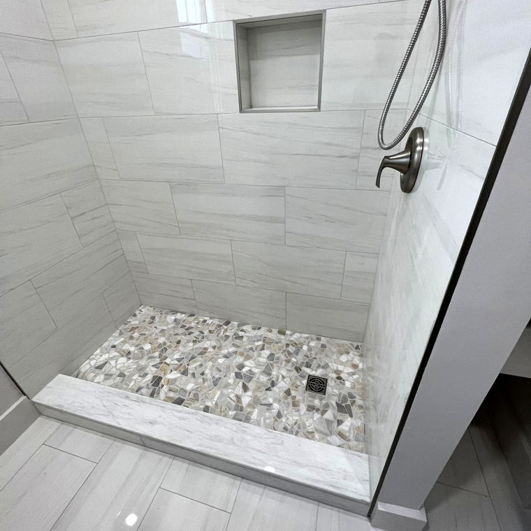 Corner of a grey striated porcelain shower showing the pebble mosaic pan, marble curb and the floor beyond