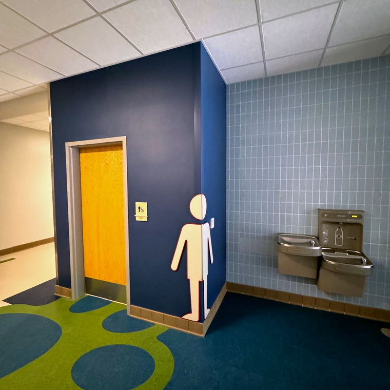 Restroom entrance with a navy wall and figure graphic beside a pale blue tiled drinking fountain alcove