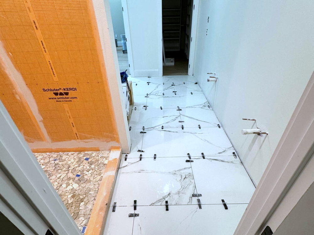 Shower wall covered in orange Schluter KERDI waterproofing membrane beside a marble-look porcelain floor being set with leveling clips