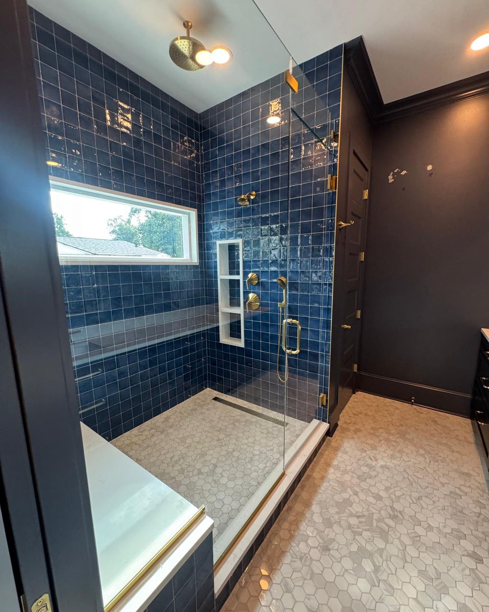 Shower lined in deep navy glazed square tile with brushed brass fixtures, a quartz bench, a three-shelf niche and a marble hexagon floor