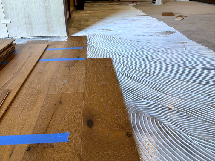 Close view of wide-plank oak flooring being glued down, with adhesive combed in even ridges by a notched trowel