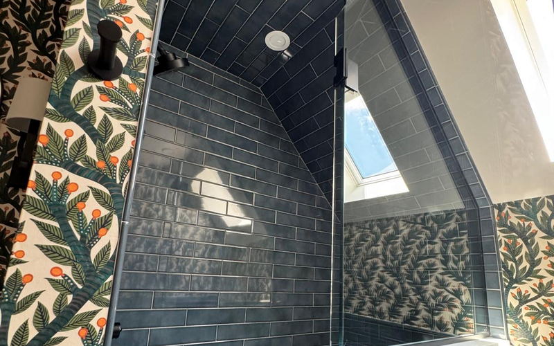 Shower lined in dark blue subway tile following a sloped dormer ceiling, with frameless glass and botanical wallpaper beyond